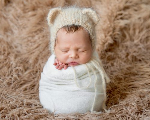 Adorable newborn baby in a beige bonnet swaddled in a white wrap sleeping tightly on furry surface.