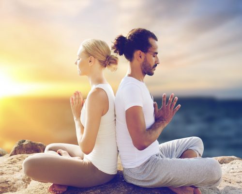 yoga , mindfulness, harmony and people concept - happy couple meditating in lotus pose outdoors over sea background